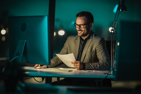 Male businessmen in a grey suit staring at a computer screen while holding a blank piece of paper at night in the office