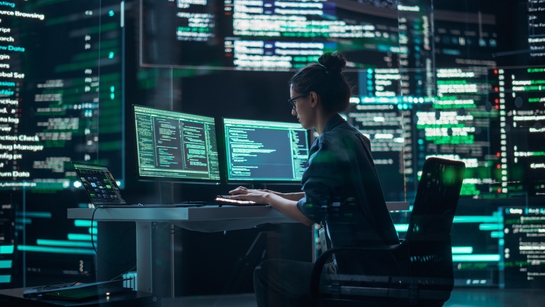 A roofing female expert reviewing data over a dual screen monitor in an office at night