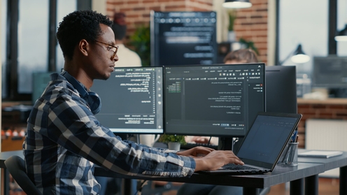 A male developer working at a desk with two monitors and a laptop, focused on parsing algorithm in software agency in a modern office setting