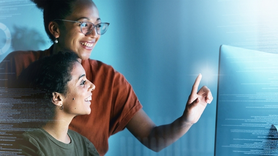 Two female retail employees smiling and reviewing lines of code in an office at night