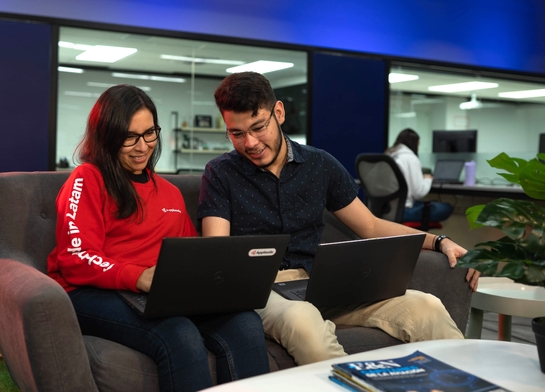 Two Applaudo software developers seated on a couch, each using a laptop, engaged in a focused discussion or work session.