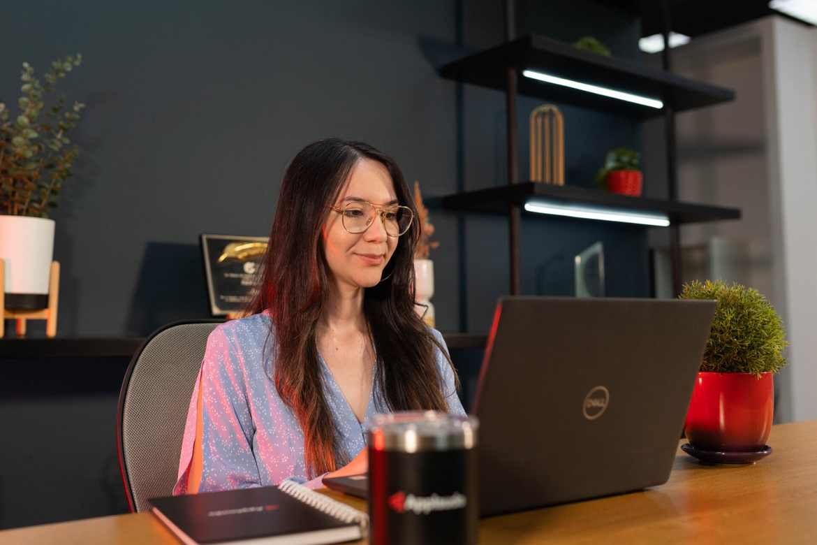 A woman seated at a desk, focused on her laptop, surrounded by a tidy workspace.